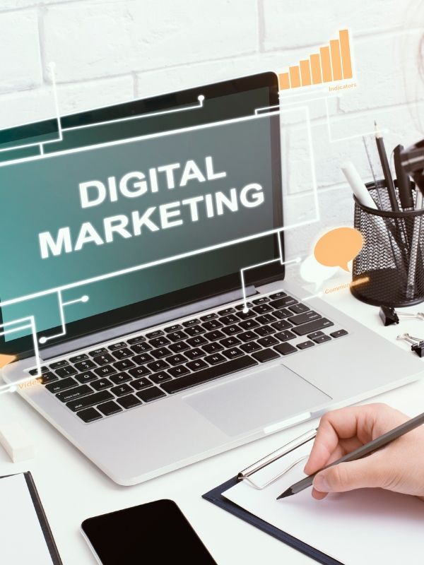 A laptop showing the words digital marketing on a desk beside a hand taking notes